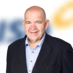 Bald man in a dark blazer and light blue shirt, smiling at the camera on a blurred background.