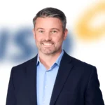 Smiling man in a navy suit and light blue shirt, standing in front of a blurred white background.