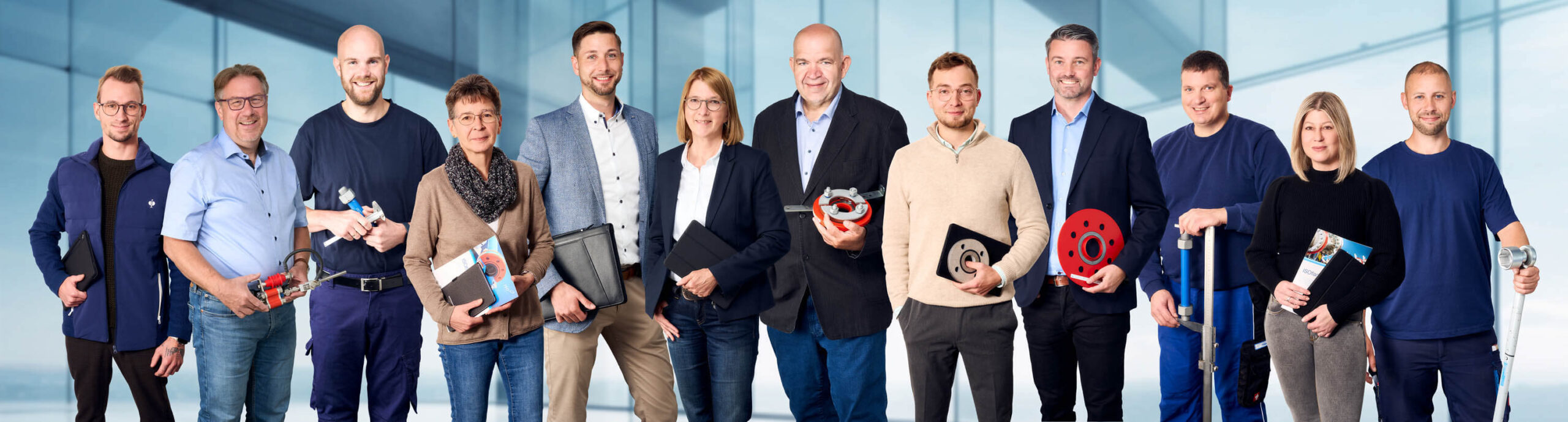 Twelve people stand in a row, smiling, some holding tools, folders, or mechanical parts in an office setting.